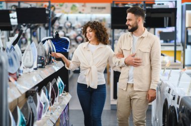 Çift bir elektronik mağazasında çeşitli ev aletlerini inceler, seçenekleri tartışır ve satın almaları için özellikleri karşılaştırır
