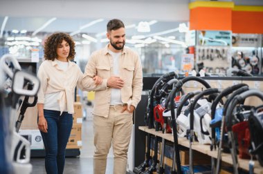 Genç çift, çeşitli seçeneklerle çevrili canlı bir elektronik mağazasında birlikte alışveriş yaparken yeni bir elektrikli süpürge seçmekten mutluluk duyuyorlar.