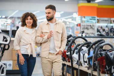 Genç çift bir elektronik mağazasına göz gezdiriyorlar ve satın alma kararı alırken çeşitli elektrik süpürgesi seçeneklerini tartışıyor ve karşılaştırıyorlar.