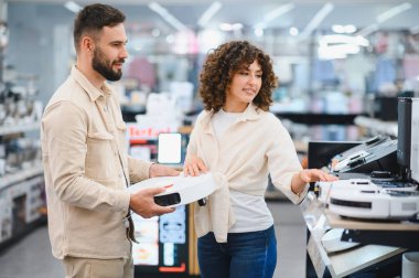Robot elektrik süpürgelerini karşılaştıran genç bir çift modern elektronik mağazasından akıllı ev aletleri alışverişi yapıyor.