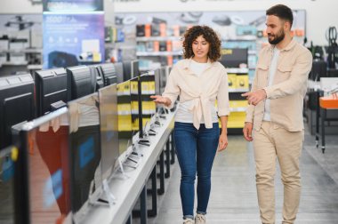 Genç çift bir tüketici elektroniği mağazasına göz gezdirirken, ekrandaki çeşitli televizyonları karşılaştırırken seçenekleri tartışıyor