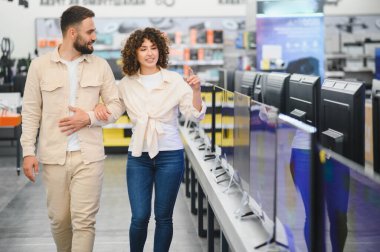 Genç bir çift elektronik mağazasında geziniyor, ev eğlencesi için yeni bir televizyon seçiyor ve inceliyor.