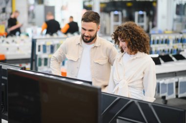 Genç çift, bir tüketici elektroniği mağazasında gezinirken, ev eğlenceleri için en son televizyonları düşünürken seçenekleri karşılaştırıyor
