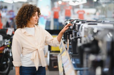 Müşteri, modern teknoloji seçenekleriyle çevrili, hareketli bir mağazada en iyi anlaşmayı ararken çeşitli kahve makinelerini karşılaştırıyor.