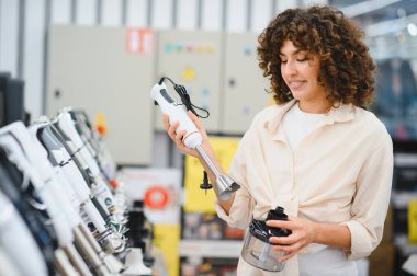 Bir elektronik mağazasında farklı daldırma mikseri modellerini karşılaştıran genç bir kadın. Bir tanesini elinde tutuyor ve dikkatlice bakıyor.