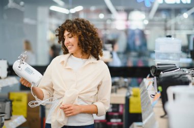 Gülümseyen kadın ev aletleri mağazasında alışveriş yaparken el mikseri seçiyor, çeşitli mutfak gereçleri seçeneklerinin tadını çıkarıyor.