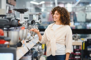 Gülümseyen kadın perakende mağazasında ev aletlerini seçiyor, çeşitli yemek işlemcileri ve et öğütücülerini inceliyor.