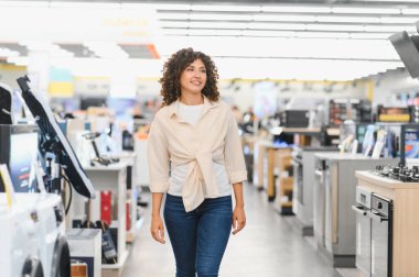 Gülümseyen kadın elektronik mağazasında yürüyor ev aletlerini seçiyor, çeşitli ürünlere bakıyor.