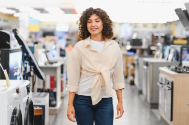 Genç bir kadın elektronik mağazasına giriyor, çeşitli ev aletlerine bakıyor ve ne alacağına karar veriyor.