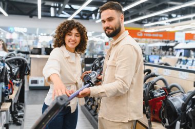 Genç çift ev aletleri mağazasına göz gezdiriyorlar, çeşitli seçenekleri ve modelleri düşünürken yeni bir elektrikli süpürge seçiyorlar.