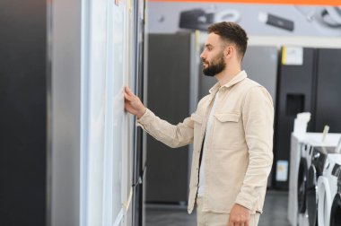 Sakallı bir adam, bir mağazadaki çeşitli buzdolabını karıştırıyor ve mutfak güncellemesi için mükemmel modeli dikkatlice seçiyor.