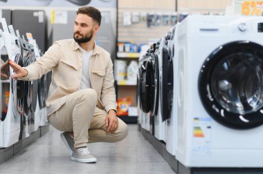 Sakallı adam yeni bir çamaşır makinesini incelerken bir ev aletleri mağazasında alışveriş yapıyor seçenekleri karşılaştırıyor ve seçimler yapıyor.