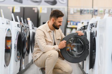 Sakallı bir adam yeni bir çamaşır makinesini inceliyor. Modern ev aletleri alırken seçenekleri dikkatlice değerlendiriyor.