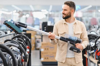 Sakallı bir adam elektronik mağazasında kablosuz elektrik süpürgesi tutarken ev aletlerini karıştırıyor.