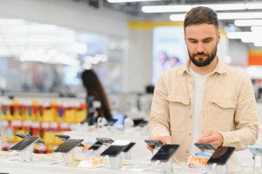 Sakallı adam yeni akıllı telefonları karşılaştırırken en son teknoloji gereçleriyle dolu bir mağazadan en iyi cep telefonunu seçiyor.