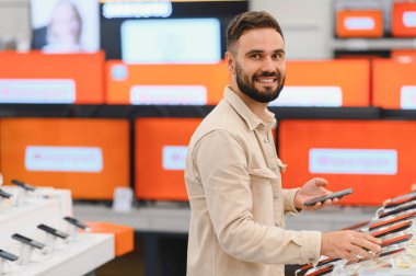 Tüketici elektroniği mağazasında alışveriş yaparken yeni bir akıllı telefonu karşılaştıran ve seçen sakallı bir adam, seçenekleri keşfetme deneyiminin tadını çıkarıyor