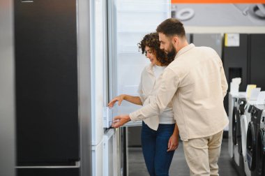 Genç çift bir elektronik mağazasında buzdolabını dikkatle inceliyor ve ev aletlerinin alımı hakkında bilgilendirilmiş kararlar veriyorlar.