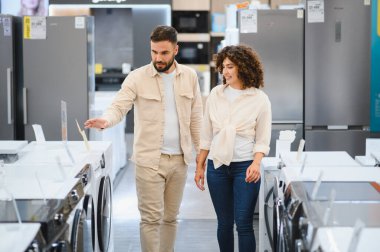 Genç çift ev aletleri mağazasından yeni çamaşır makinesi seçiyor. Satın almadan önce modelleri ve özellikleri karşılaştırıyorlar.