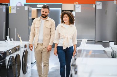 Genç bir çift, elektronik mağazasında sıra sıra çamaşır makinelerinde dolaşıp evleri için mükemmel aletleri arıyorlar.
