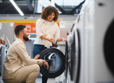 Elektronik mağazasında gülümseyen bir çift çamaşır makinesini inceliyor, ev aletleri almaya karar veriyorlar.