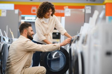 Çift bir elektronik mağazasında çamaşır makinelerini inceliyor, ev aletlerinin alımı için özellikleri ve seçenekleri tartışıyor