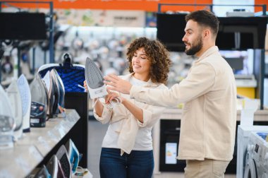 Elektronik mağazasında yeni bir ütü seçen genç çift, satın almadan önce farklı modelleri inceliyor.