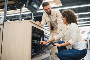 Ev aletleri mağazasında yeni bir fırın seçen mutlu çift, kadın fırının kapısını açıyor ve ortağına gülümsüyor.