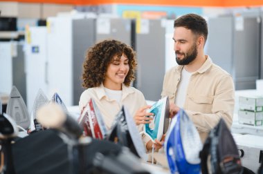 Genç çift yeni buhar ütüsünü seçerken gülümsüyor. Parlak ve modern bir ev aletleri mağazasında, alışveriş deneyimlerinin tadını çıkarıyorlar.
