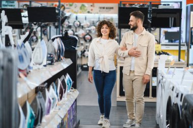 Bir elektronik mağazasında kol kola yürüyen mutlu çift, birlikte eğlenirken ev aletlerini seçiyorlar.
