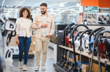 Elektronik mağazasında kol kola yürüyen mutlu çift, birlikte yeni bir elektrikli süpürge seçiyorlar, alışveriş deneyiminin tadını çıkarıyorlar.