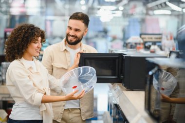 Mutlu çift mikrodalga fırının boyutunu kontrol ediyor, plastik konteynır tutuyor, elektronik mağazasından ev aletleri alıyor.