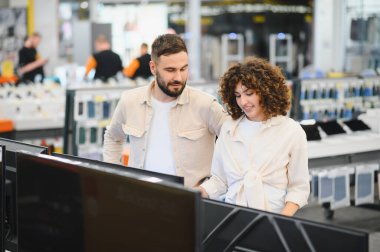 Genç çift, tüketici elektroniği mağazasında yeni bir televizyon arıyor. Seçenekleri karşılaştırıyor ve alışveriş deneyimlerinin keyfini çıkarıyorlar.