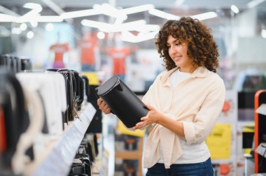 Elektronik mağazasından siyah elektrikli su ısıtıcısını seçen gülümseyen kadın, cihazı tutuyor ve kutudaki bilgileri okuyor.