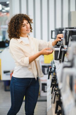 Bir elektronik mağazasında ev aletleri alırken çeşitli kahve makinelerini karşılaştıran genç bir kadın, seçenekleri ve özellikleri değerlendiriyor