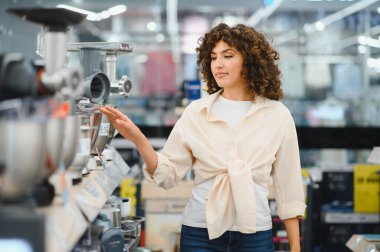 Genç bir kadın mutfak aletlerini seçerken bir et öğütücüsünü işaret ediyor. Parlak, düzenli bir elektronik mağazası koridorunda.