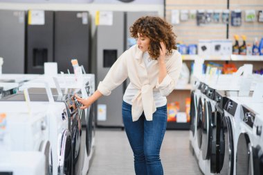 Müşteri, ev aletleri mağazasında alışveriş yaparken yeni çamaşır makinelerinin özelliklerini ve fiyatlarını karşılaştırarak satın alma seçeneklerini araştırıyor