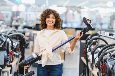 Gülümseyen kadın, elektrik süpürgesini seçerken ev aletleri almak için opsiyonlarla dolu canlı bir elektronik mağazasından alışveriş yapıyor.