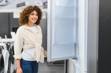 Gülümseyen kadın müşteri buzdolabı kapısını açıyor, elektronik mağazasında ev aletlerini seçiyor.