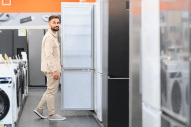 Adam buzdolabının kapısını açıp içeriyi kontrol ederken bir ev aletleri mağazasında yeni bir buzdolabı seçiyor.