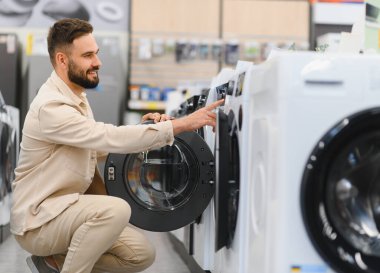 Genç adam yeni çamaşır makinesi seçiyor, özelliklerini inceliyor ve modern ev çamaşırları için satın alma kararı veriyor.