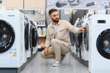 Çömelmiş genç bir adam, ev aletleri mağazasında yeni bir çamaşır makinesi seçiyor, satın almadan önce özelliklerini dikkatlice inceliyor.