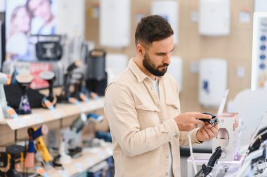Tüketici elektronik mağazasından alışveriş yaparken saç makaslarını inceleyen sakallı adam, bilgilendirilmiş bir satın alma kararı almaya odaklanmış.