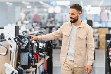 Gülümseyen müşteri, seçeneklerle dolu bir ev aletleri mağazasının koridorlarında gezinirken yeni bir elektrikli süpürge seçiyor.