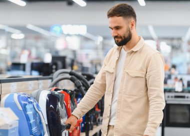 Sakallı bir adam ev aletleri mağazasından yeni bir elektrikli süpürge seçiyor, görüntüdeki farklı modelleri karşılaştırıyor.