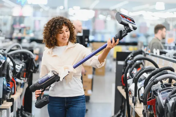 Yeni kablosuz elektrik süpürgesini seçen gülümseyen bir kadın bir elektronik mağazasında elinde tutuyor.
