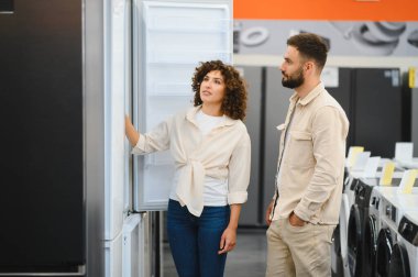 Elektronik mağazasında buzdolabını inceleyen çift ev aletleri seçeneklerini değerlendiriyor. Satın alma kararına odaklanmış görünüyorlar.