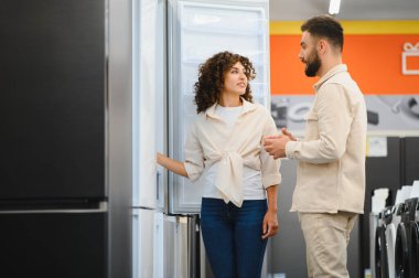 Genç çift elektronik mağazasında yeni buzdolabı seçiyor, özellikleri tartışıyor ve satın alma kararı alıyorlar.