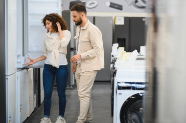 Genç bir çift ev aletleri mağazasında yeni bir buzdolabı seçiyor, seçenekleri tartışıyor ve modern mutfak ihtiyaçlarını karşılaştırıyorlar.