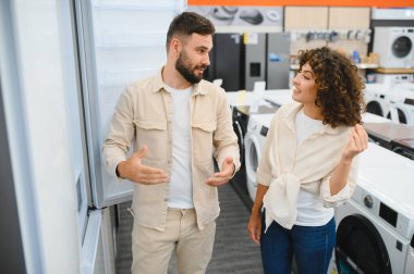 Genç çift ev aletleri mağazasında yeni buzdolabı seçiyor, özellikleri tartışıyor ve karar veriyorlar.