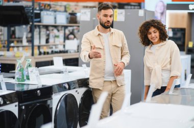 Bir çift, ev aletleri mağazasında alışveriş yaparken çeşitli çamaşır makinesi modellerini karşılaştırıyor, özellikleri tartışıyor ve birlikte karar veriyorlar.